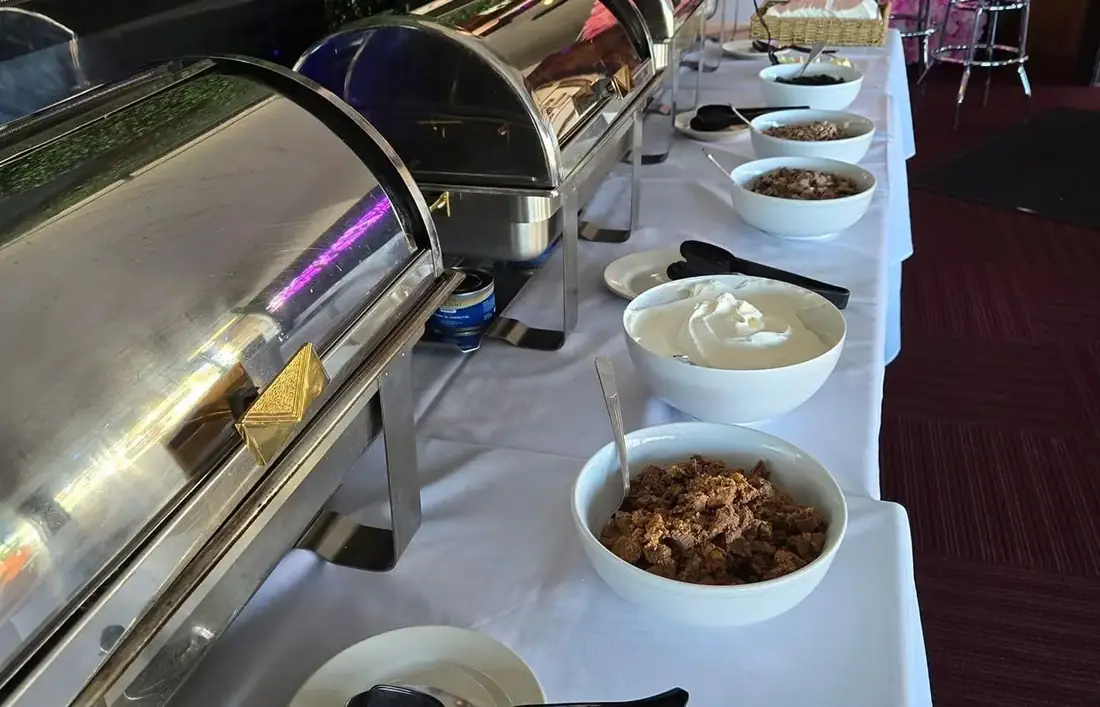 A carving station and buffet along a white-linen table beside the pink flower wall with neon angel wings.