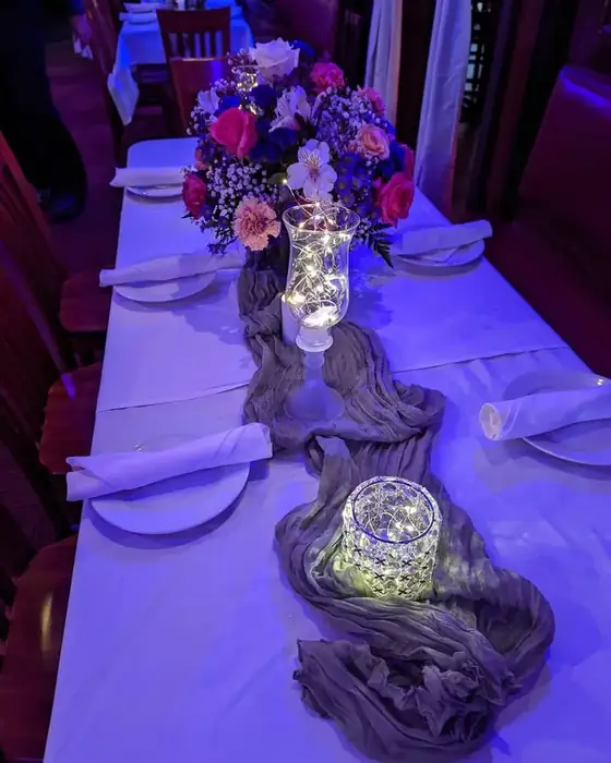 A long table styled with a cheesecloth runner, fairy lights in glass hurricanes and a floral centerpiece.